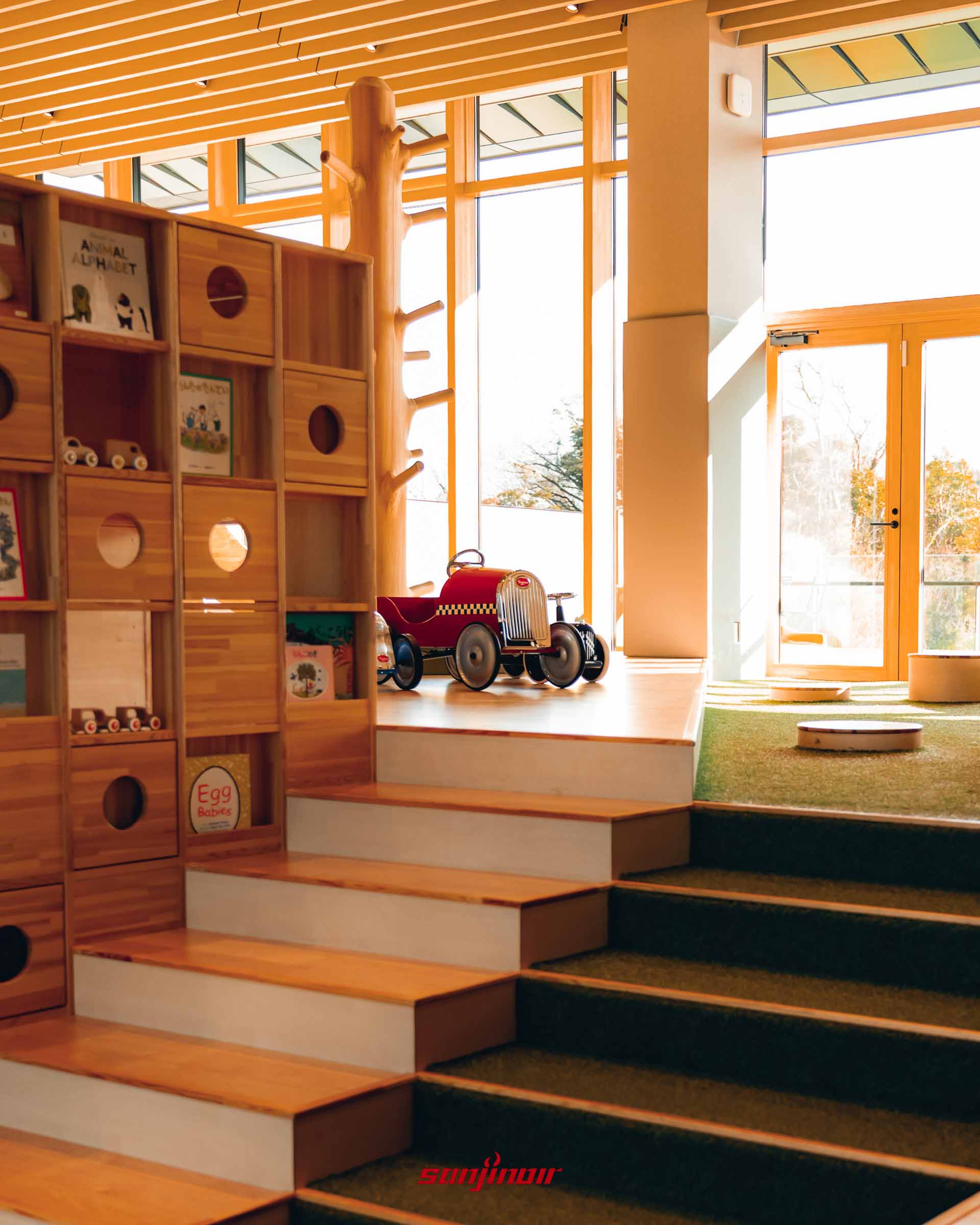 充滿木質元素的兒童遊戲區，階梯上鋪著綠色仿草皮地毯，擺放著一輛紅色復古造型的兒童踏板車與木製收納櫃。