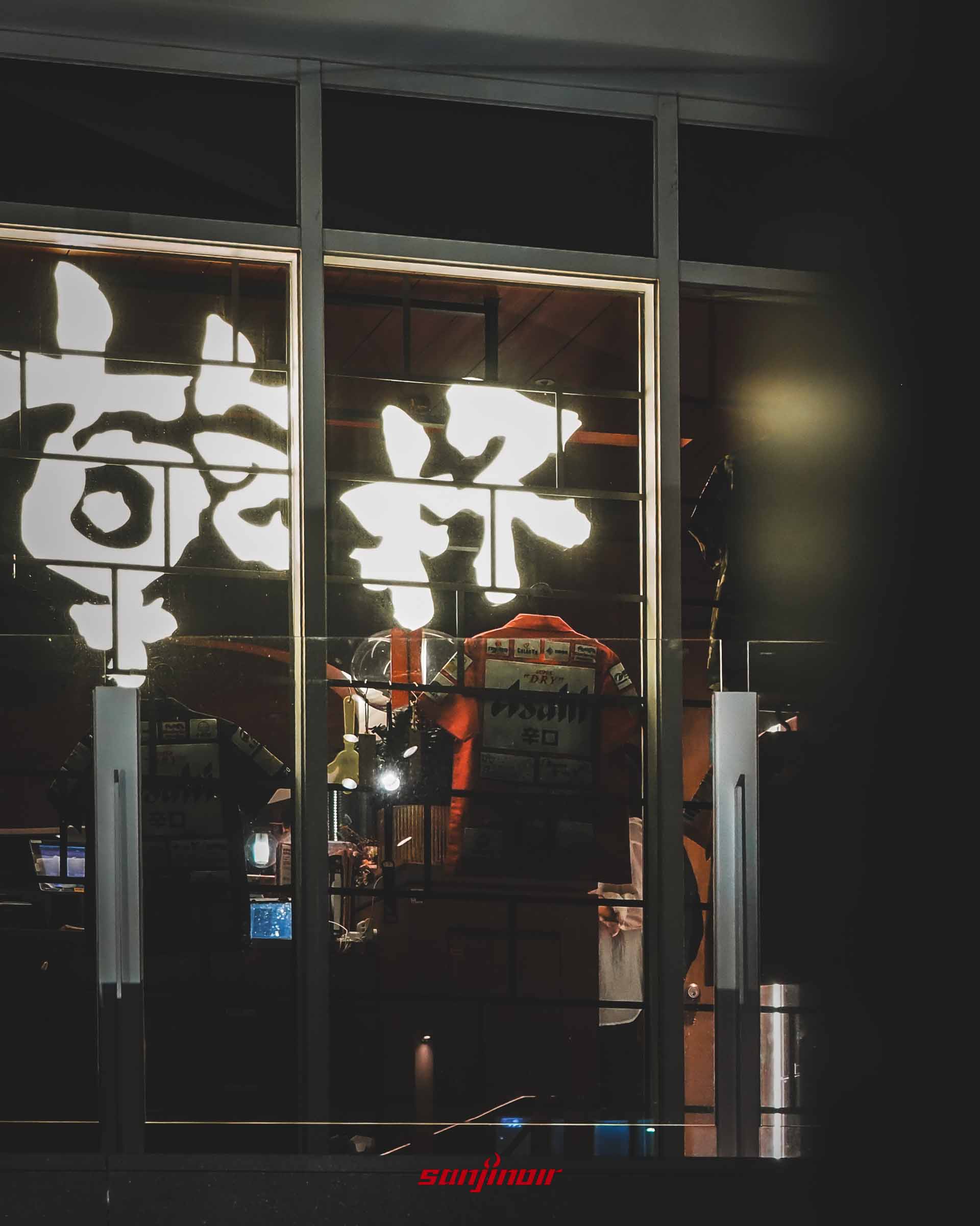 從室外看進乾杯燒肉的窗戶,玻璃上有反光的乾杯霓虹燈招牌,窗內掛著紅色Asahi啤酒制服。