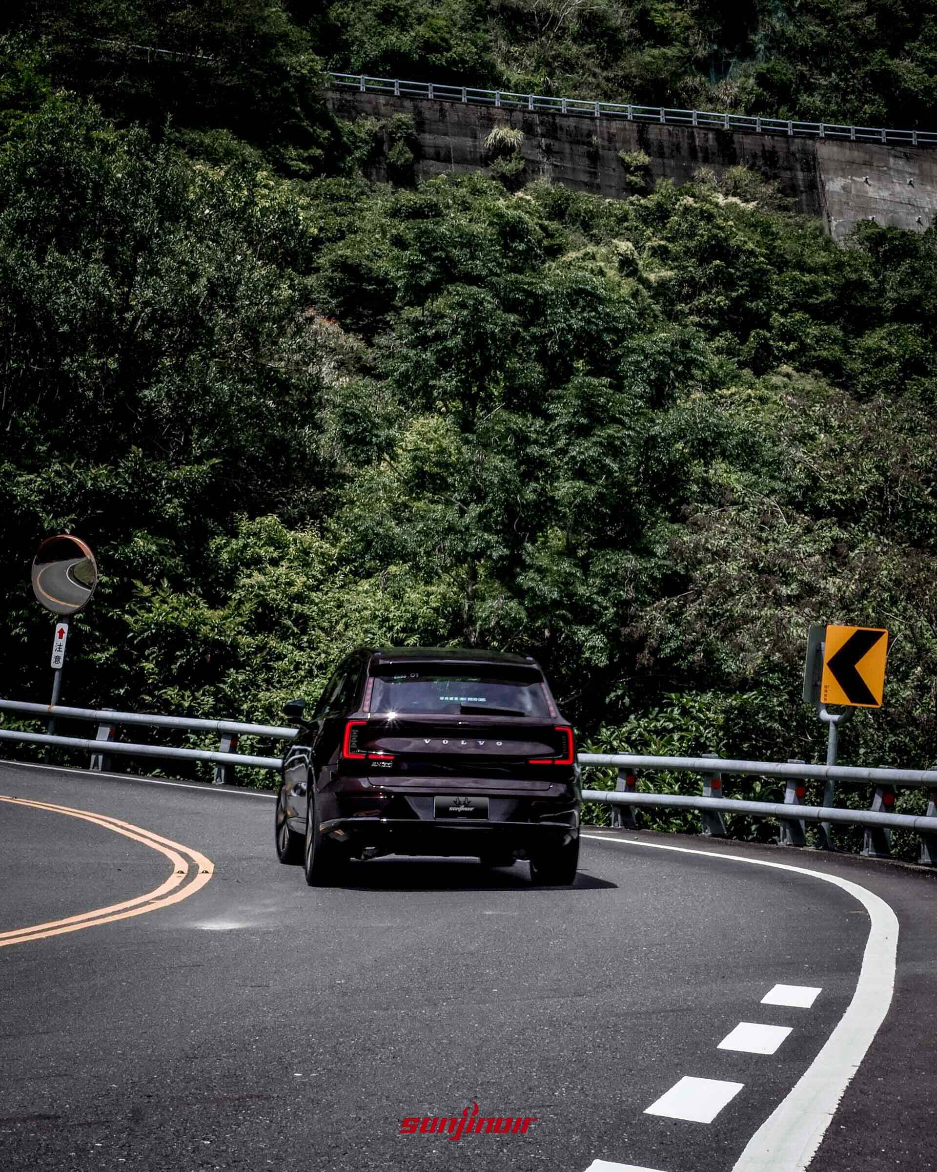 野莓紅Volvo EX90在山區右彎道路行駛的車尾動態照,後方是茂密的綠色山林與擋土牆。