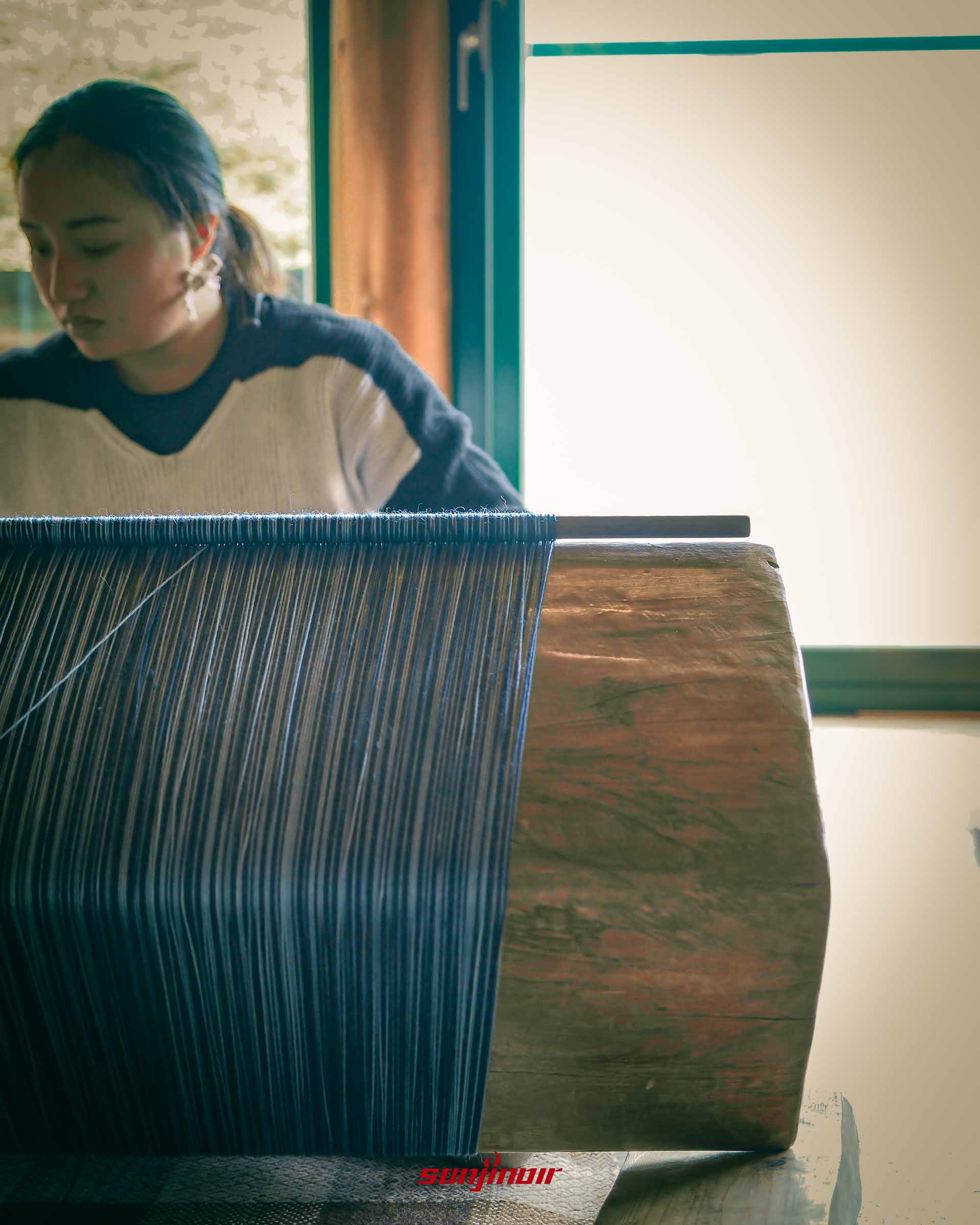 傳統織布機上密集藍色與白色絲線特寫後方為正在織布女子。