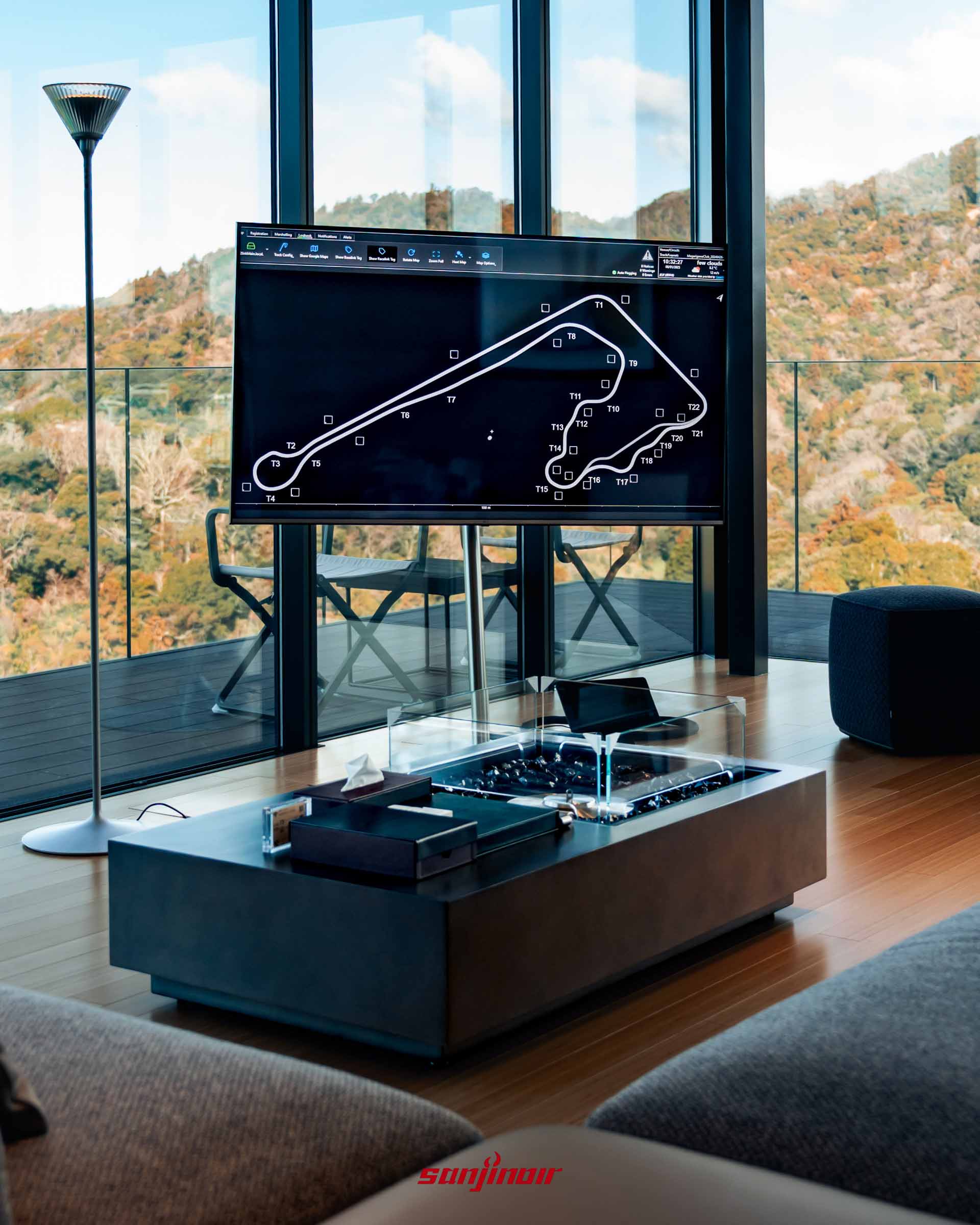 木地板客廳內設有一張中央帶有造景火爐的黑色茶几，前方電視螢幕正顯示著 MAGARIGAWA 賽道的平面路線圖。