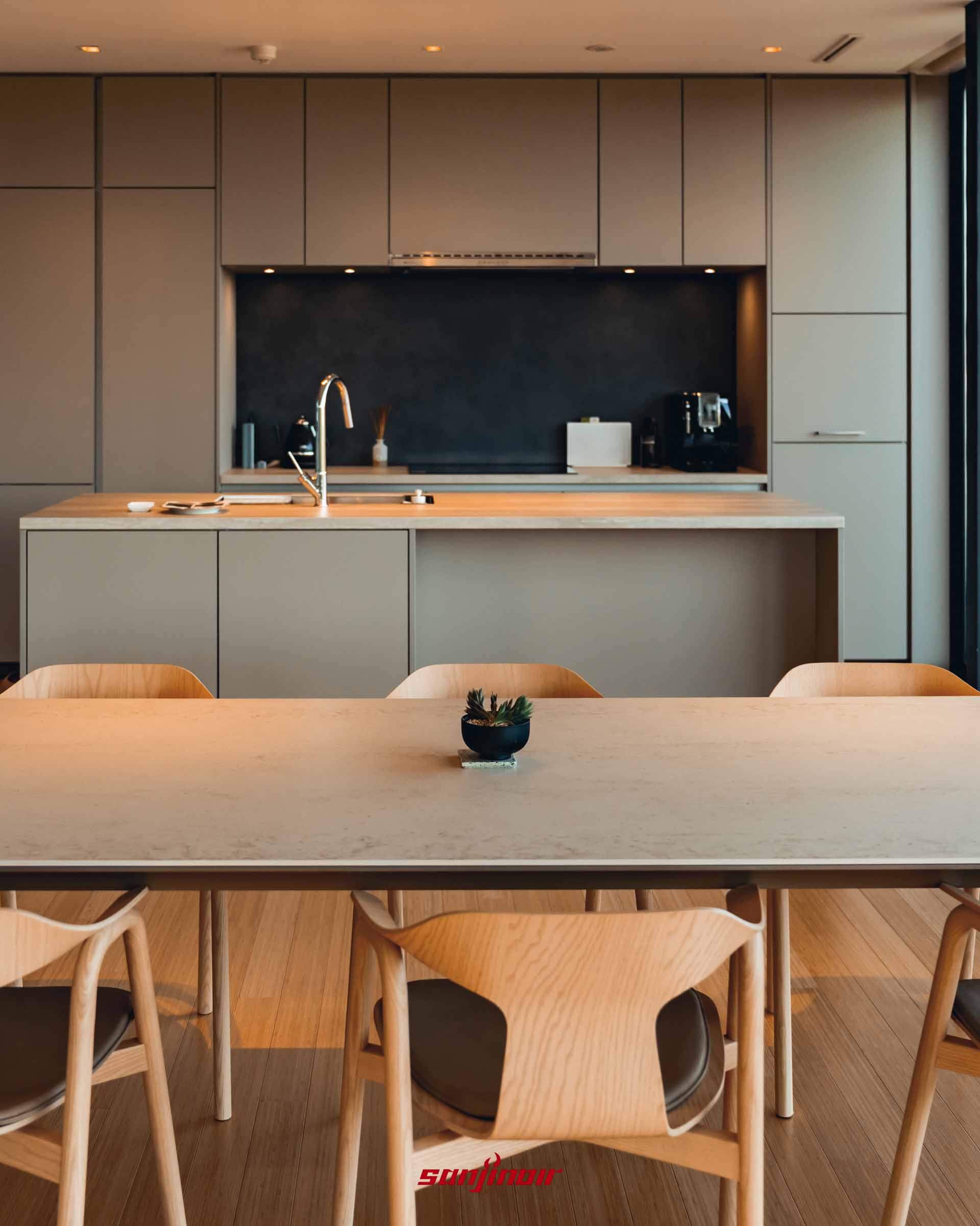 色調溫潤的開放式廚房與餐廳，前方為淺色木質大餐桌與造型單椅，後方設有寬敞的中島吧檯與深色防濺板的廚具設備。