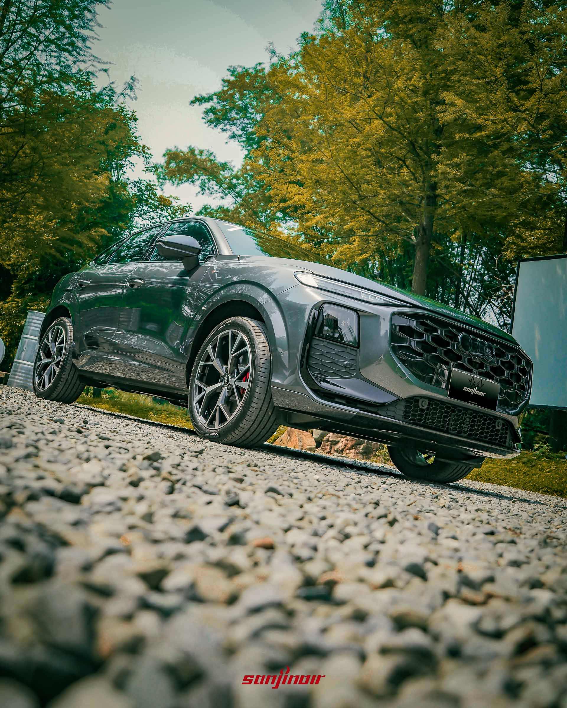 低角度拍攝Audi Q3靜止於碎石地上的全車霸氣姿態，背景為綠色樹林。