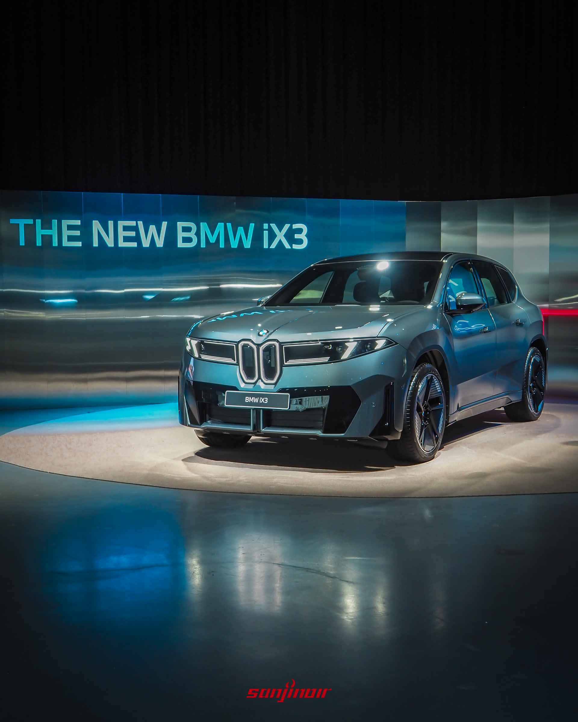 在帶有「THE NEW BMW iX3」發光字樣的藍色科技感舞台上,一輛深灰色的iX3在聚光燈下熠熠生輝。