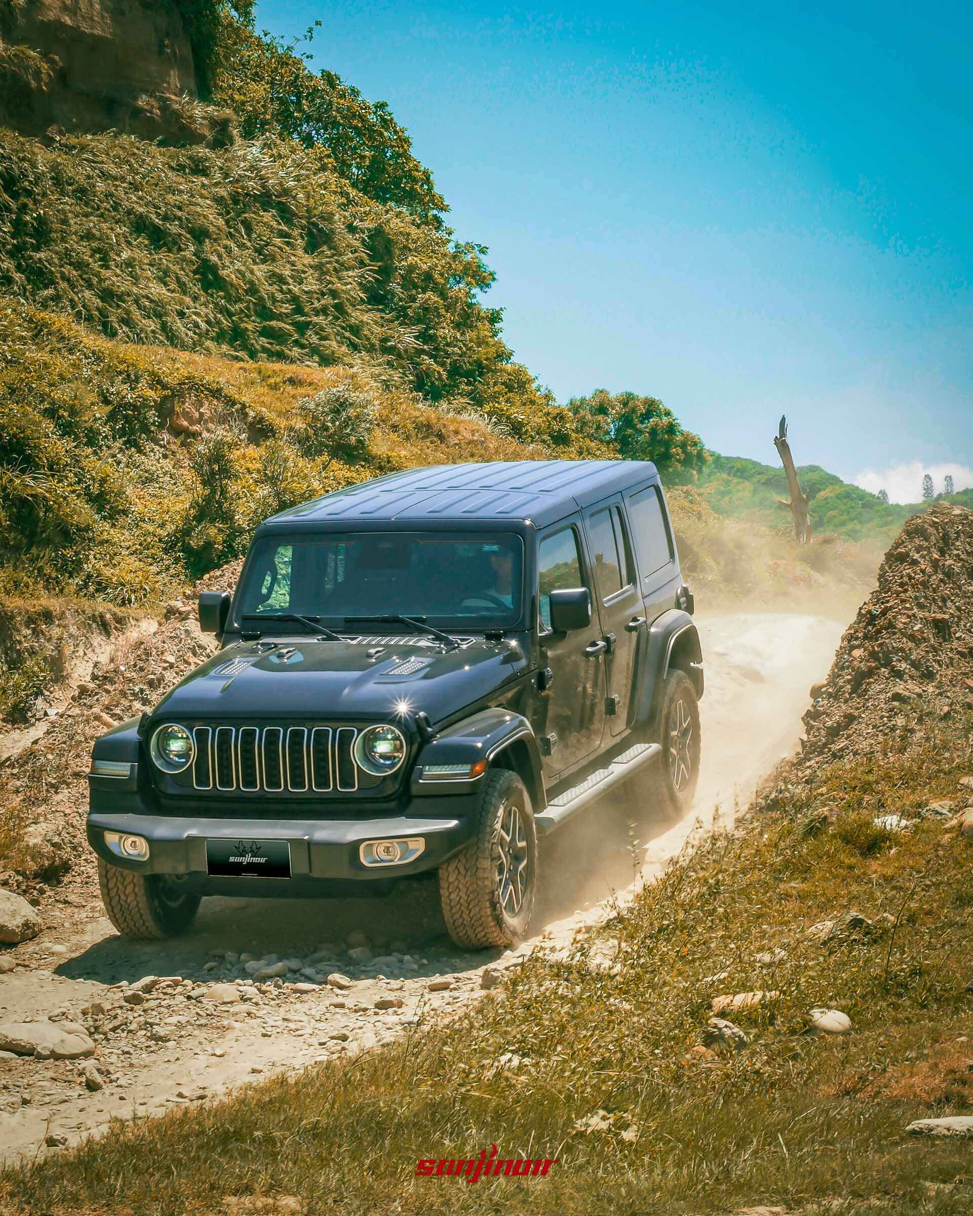 黑色 Jeep Wrangler Sahara 正在崎嶇的黃土碎石路面上行駛,揚起陣陣塵土。