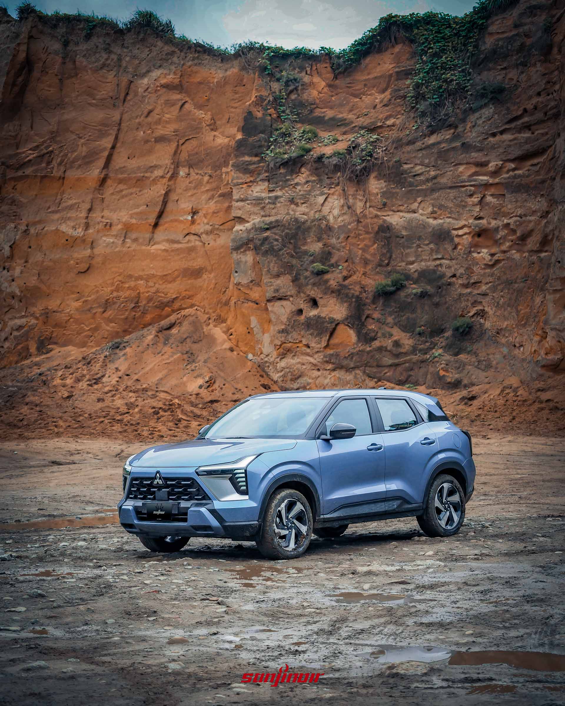藍色Mitsubishi XForce狂享版停駐在崎嶇的泥濘與礫石地面上，背景為垂直的黃褐色土崖。
