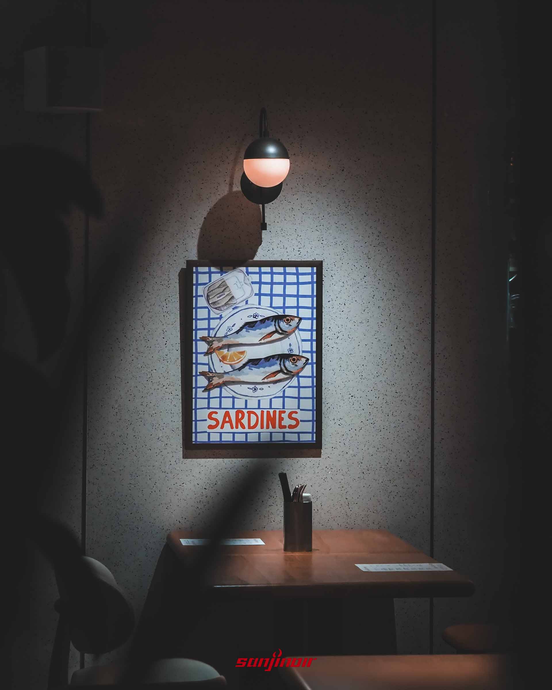 餐廳角落的雙人座位,牆上掛著一幅畫有沙丁魚的藍白格紋海報,上方設有圓形壁燈。