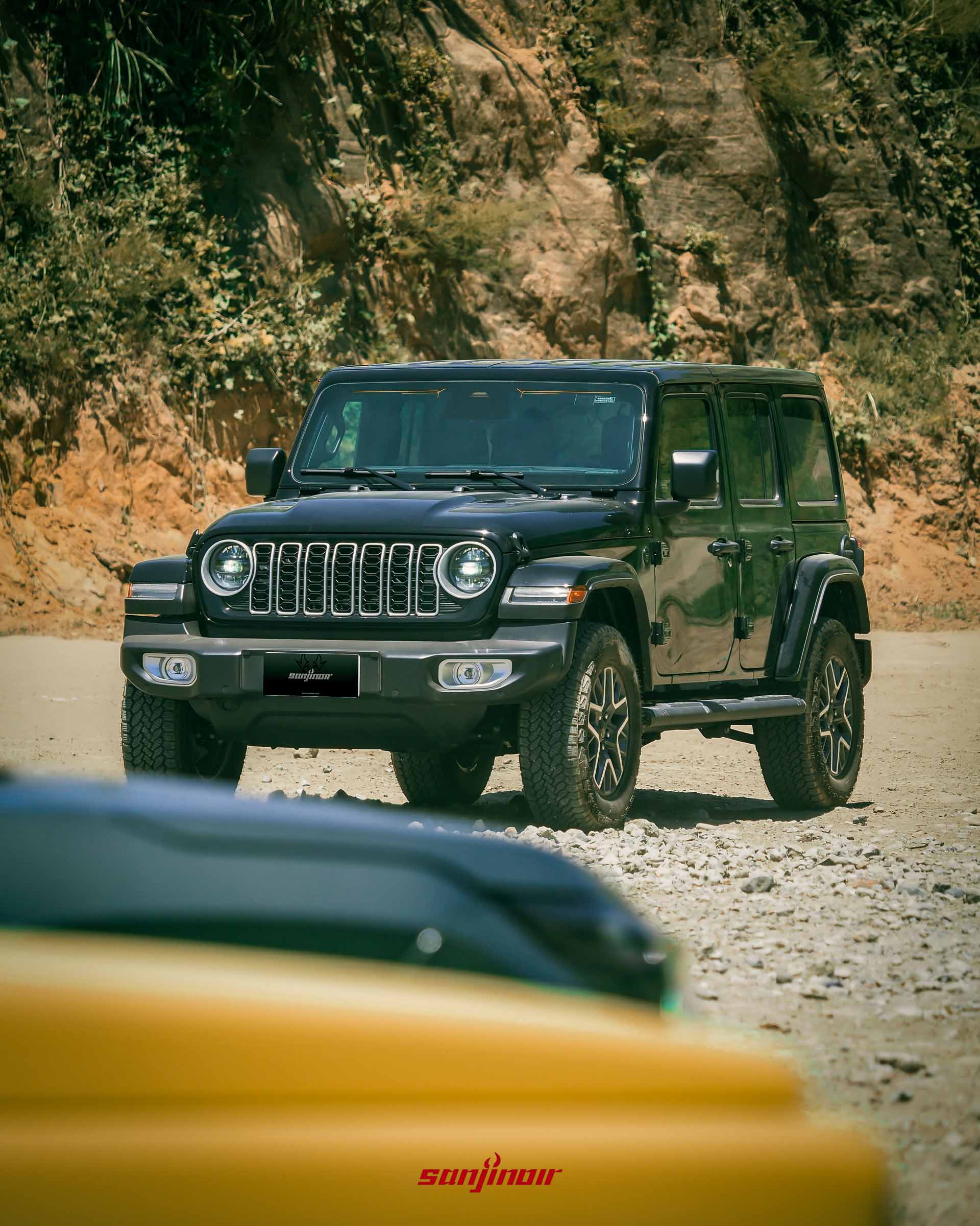 黑色 Jeep Wrangler Sahara 停在佈滿碎石的泥土空地上,背景是岩壁與綠色植被,前景有一塊黃色車頭局部入鏡。