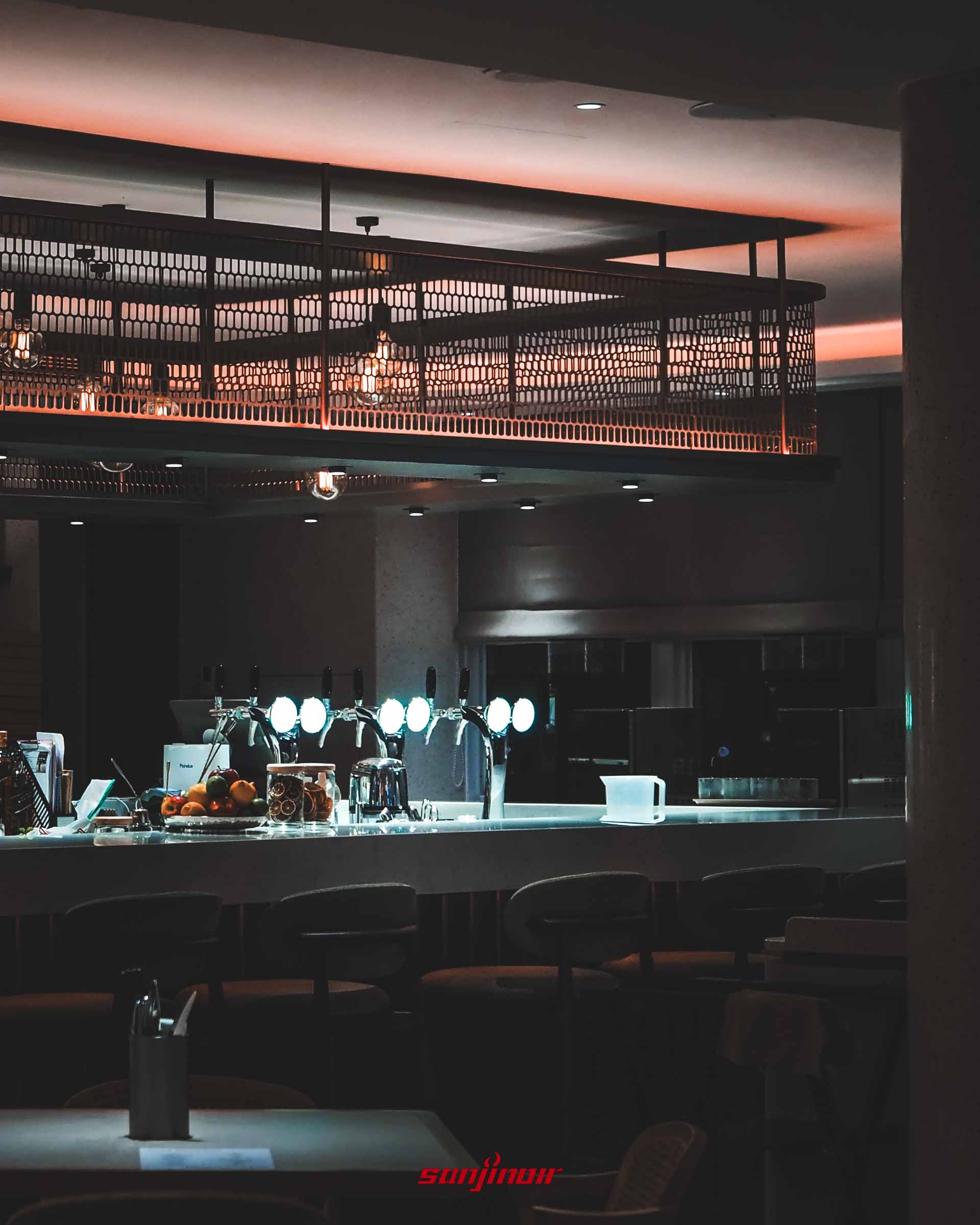 餐廳內的吧檯區,桌面上設有多支發光的生啤酒酒柱與新鮮水果,上方有金屬網狀裝飾照明。