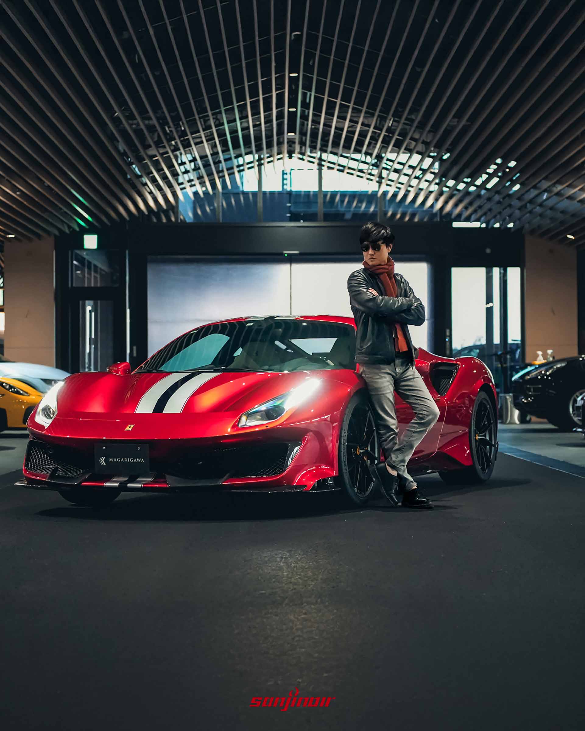 穿著皮衣配戴墨鏡的男子雙臂交叉，倚靠在紅色Ferrari 488 Pista車頭旁，背景為深灰色的頂級室內車庫空間。