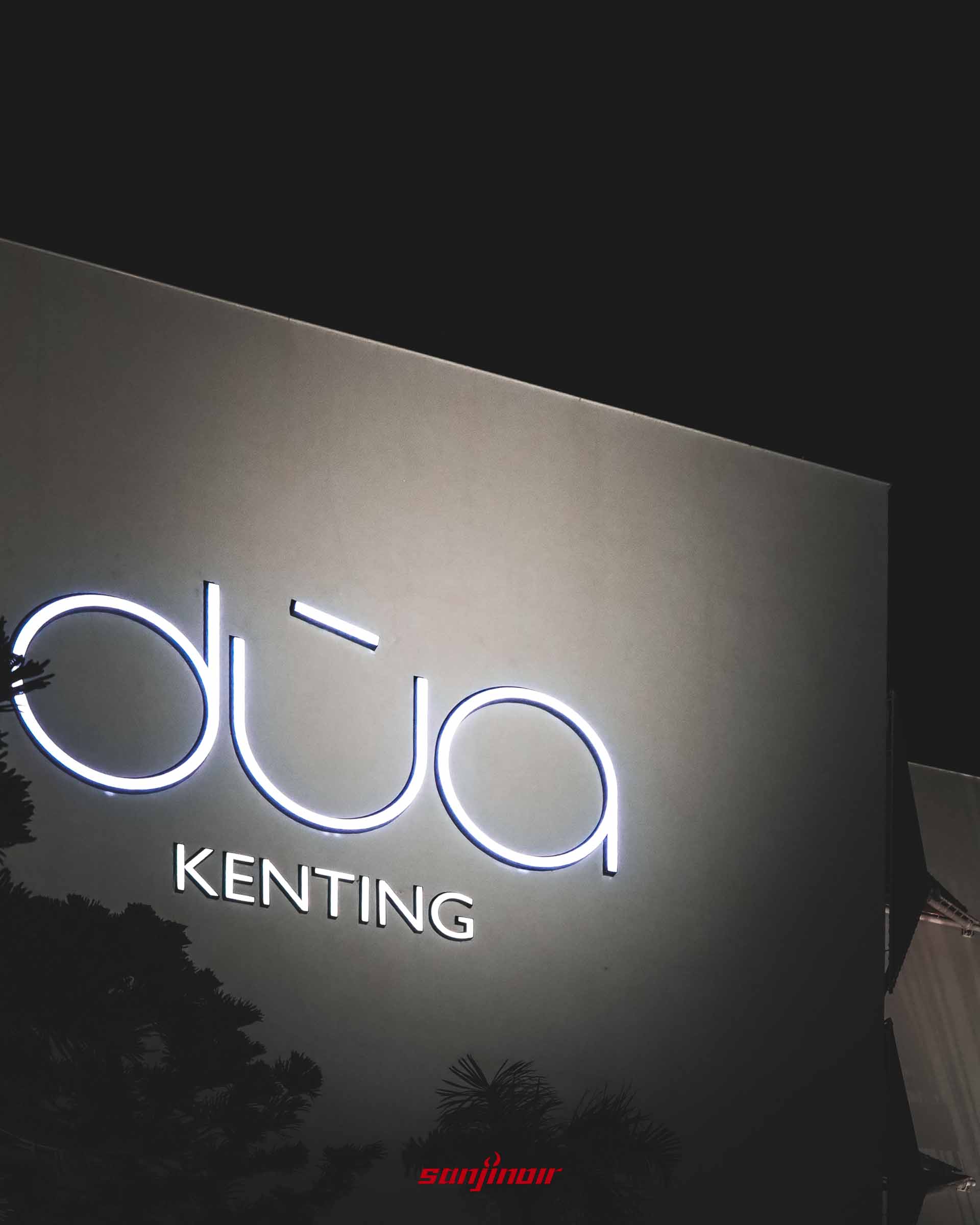 灰色牆面上的招牌近照,dùa KENTING字樣在夜色中發出白光,左下角有樹葉剪影。