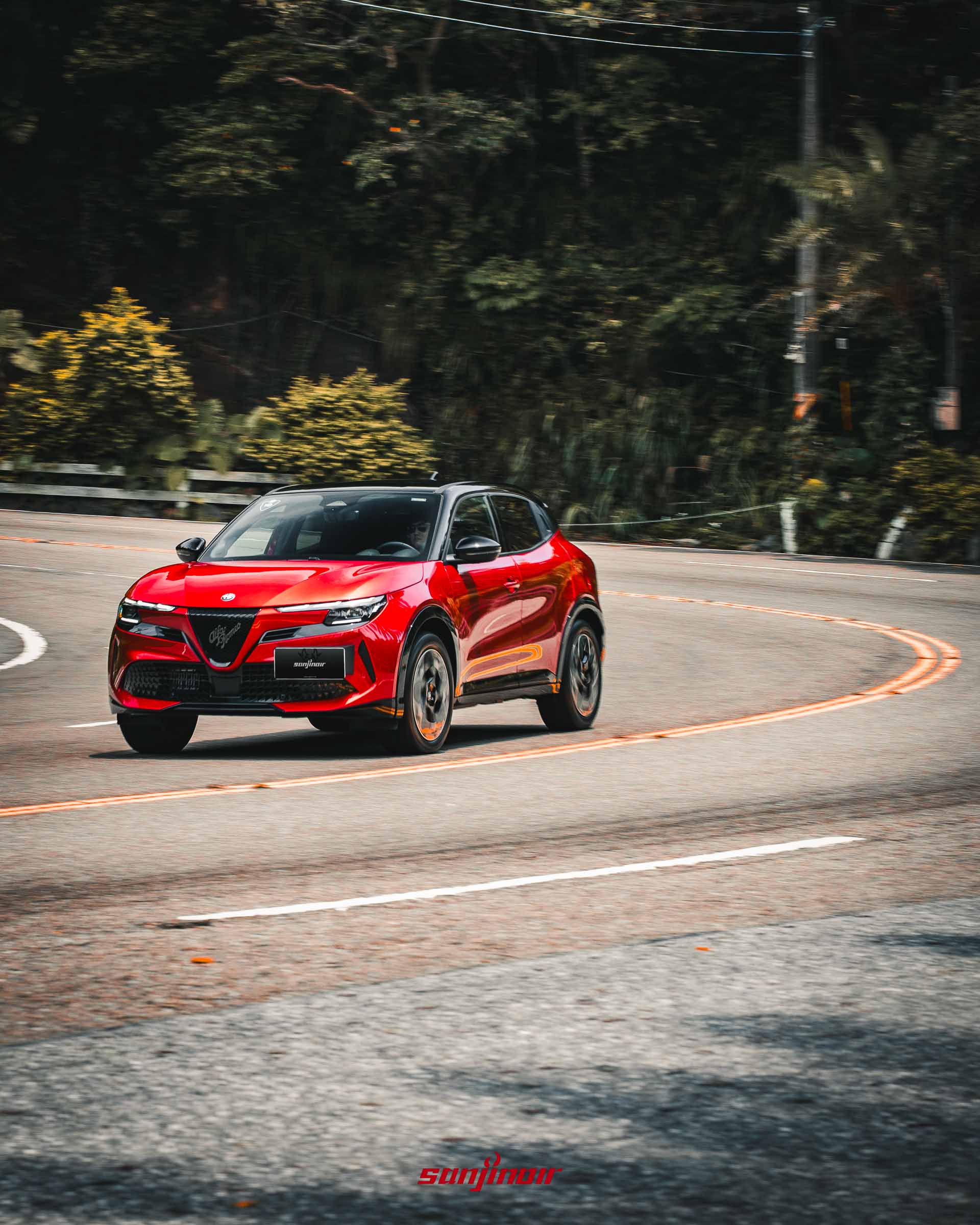 紅色Alfa Romeo Junior在山林彎道中過彎的動態追焦攝影,從右前方視角展示Scudetto水箱護罩與俐落的過彎姿態。