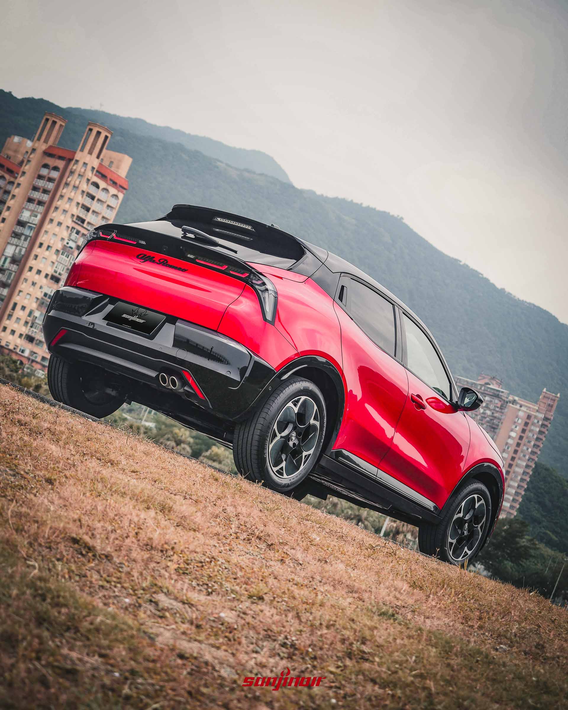從低角度拍攝停放在草坡上的紅色Alfa Romeo Junior右後方視角,背景為綿延的山丘與都會住宅大樓。