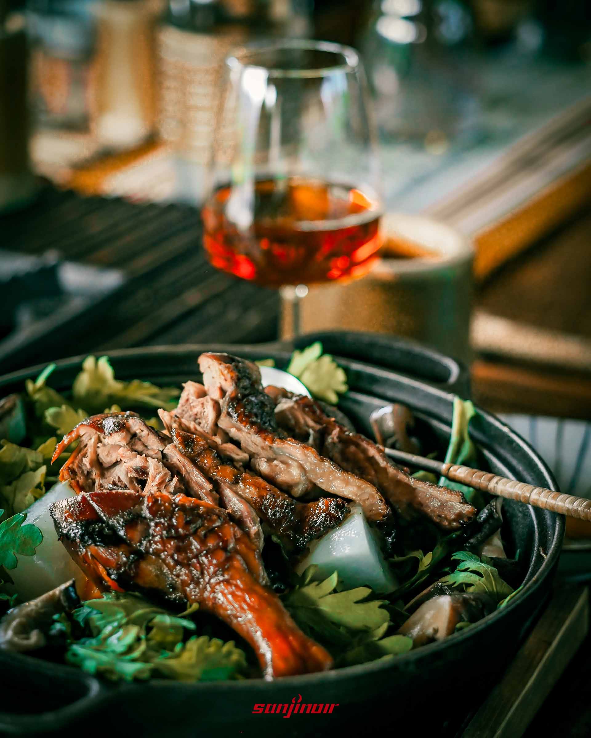 黑色鐵鍋內裝著烤鴨肉底部鋪有綠色蔬菜與白色蘿蔔後方有一杯裝在高腳杯裡的茶。