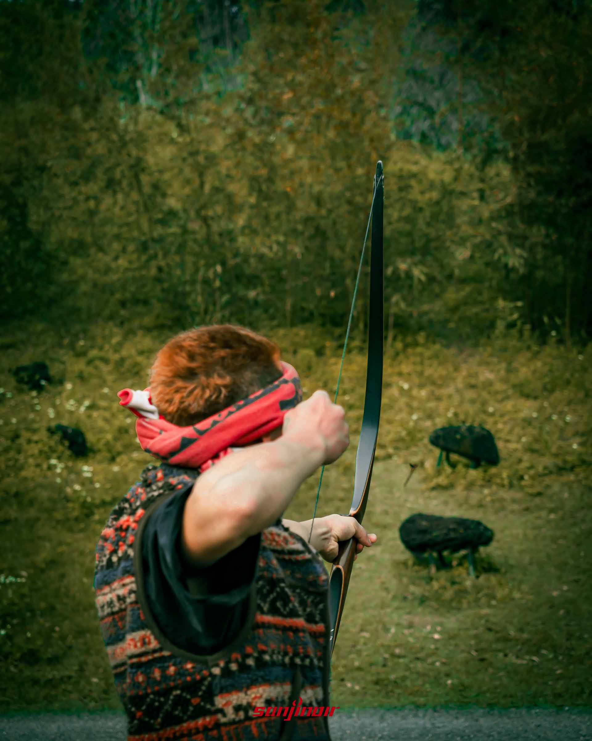 綁著紅色頭巾男子背對鏡頭拉弓準備射向草地上目標。