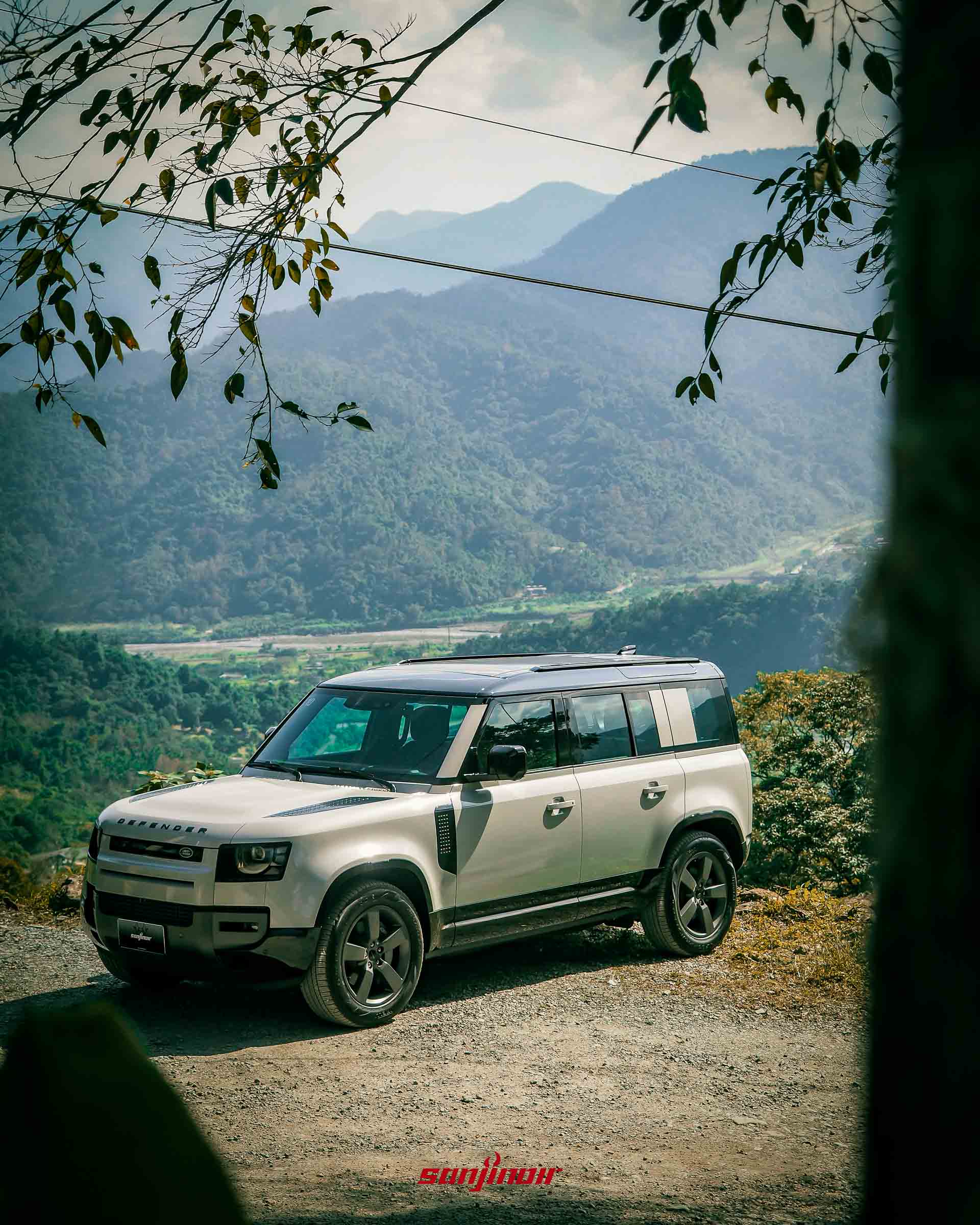 銀色Defender停靠於碎石空地背景為壯麗層疊山巒與藍天。