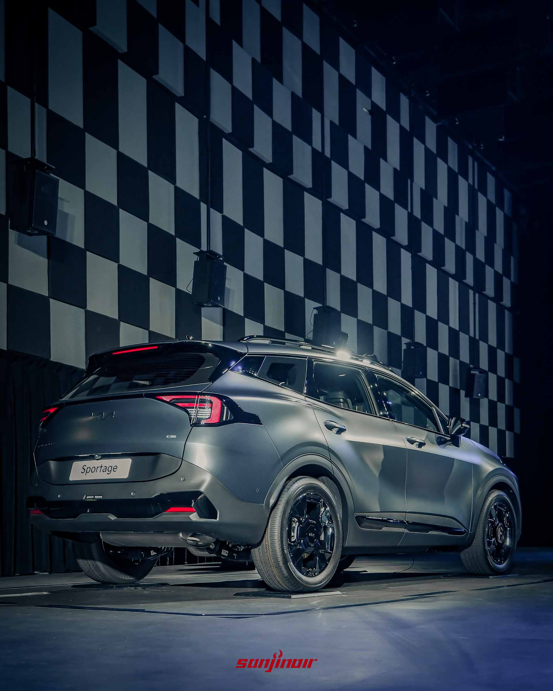 從後45度角拍攝一台停在室內舞台上的消光夜隱灰Kia Sportage X-Line休旅車。車尾LED燈點亮，展示Sportage銘板、X-line銘板。車身配備19吋黑化鋁圈、黑化車頂行李架、黑化後視鏡與車側黑化飾板。背景為黑白格子牆與黑色窗簾。